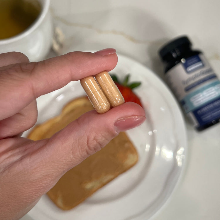 Hand holding two capsules with a plate of food and a bottle in the background